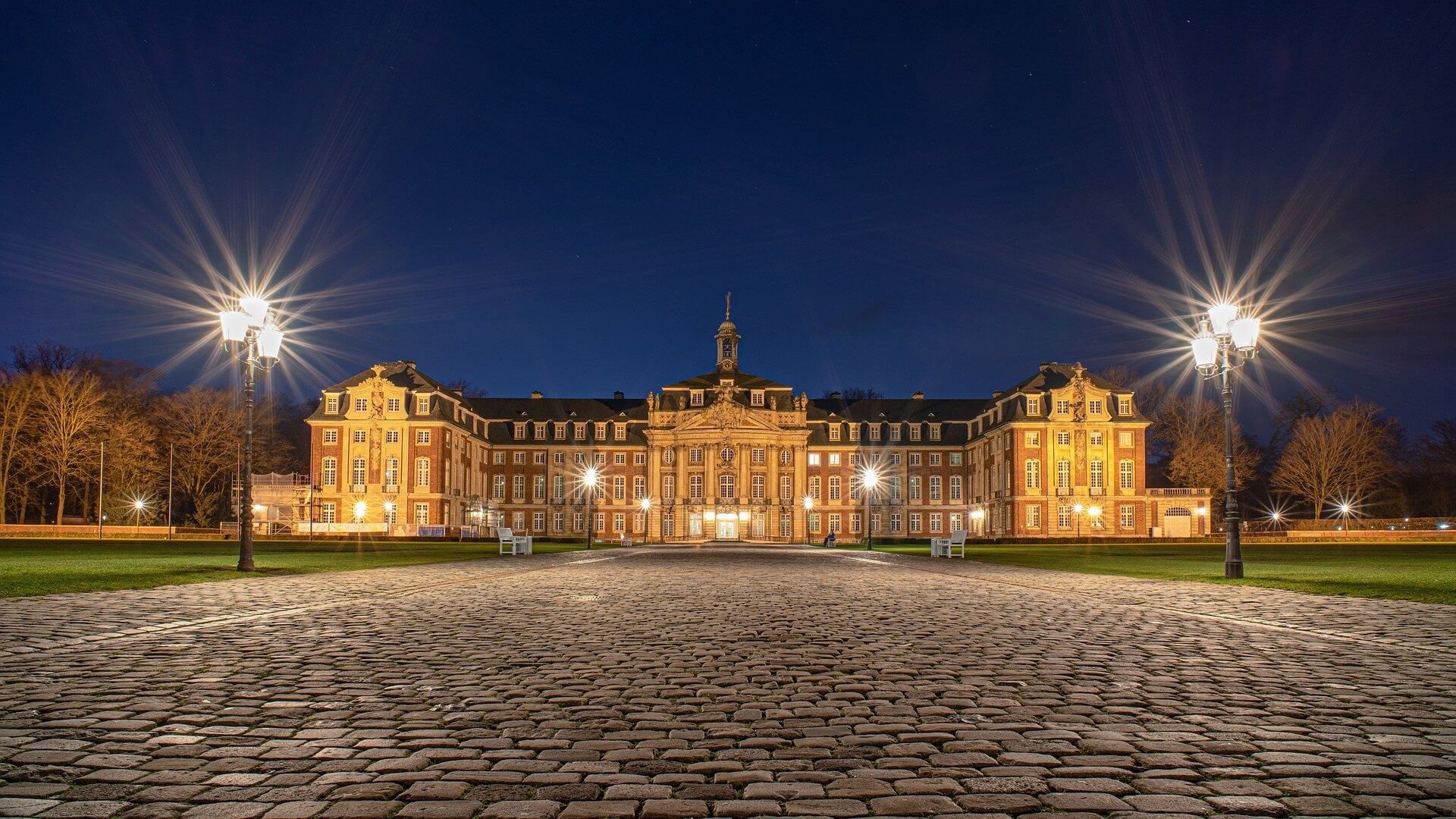 Beleuchtetes barockes Schloss mit symmetrischem Aufbau, zentralem Turm und zwei Straßenlaternen im Vordergrund bei Nacht.